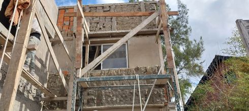 Andamios de madera alrededor de una casa de ladrillo en construcción, con árboles y un tejado al fondo.
