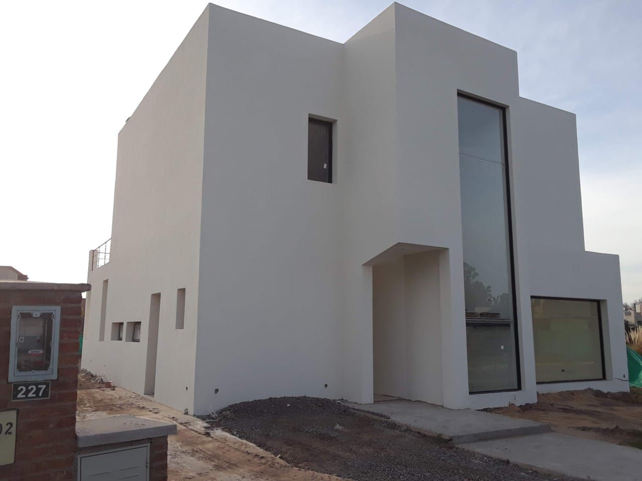 Casa moderna blanca de dos plantas en construcción con ventanas altas y patio delantero sin terminar.
