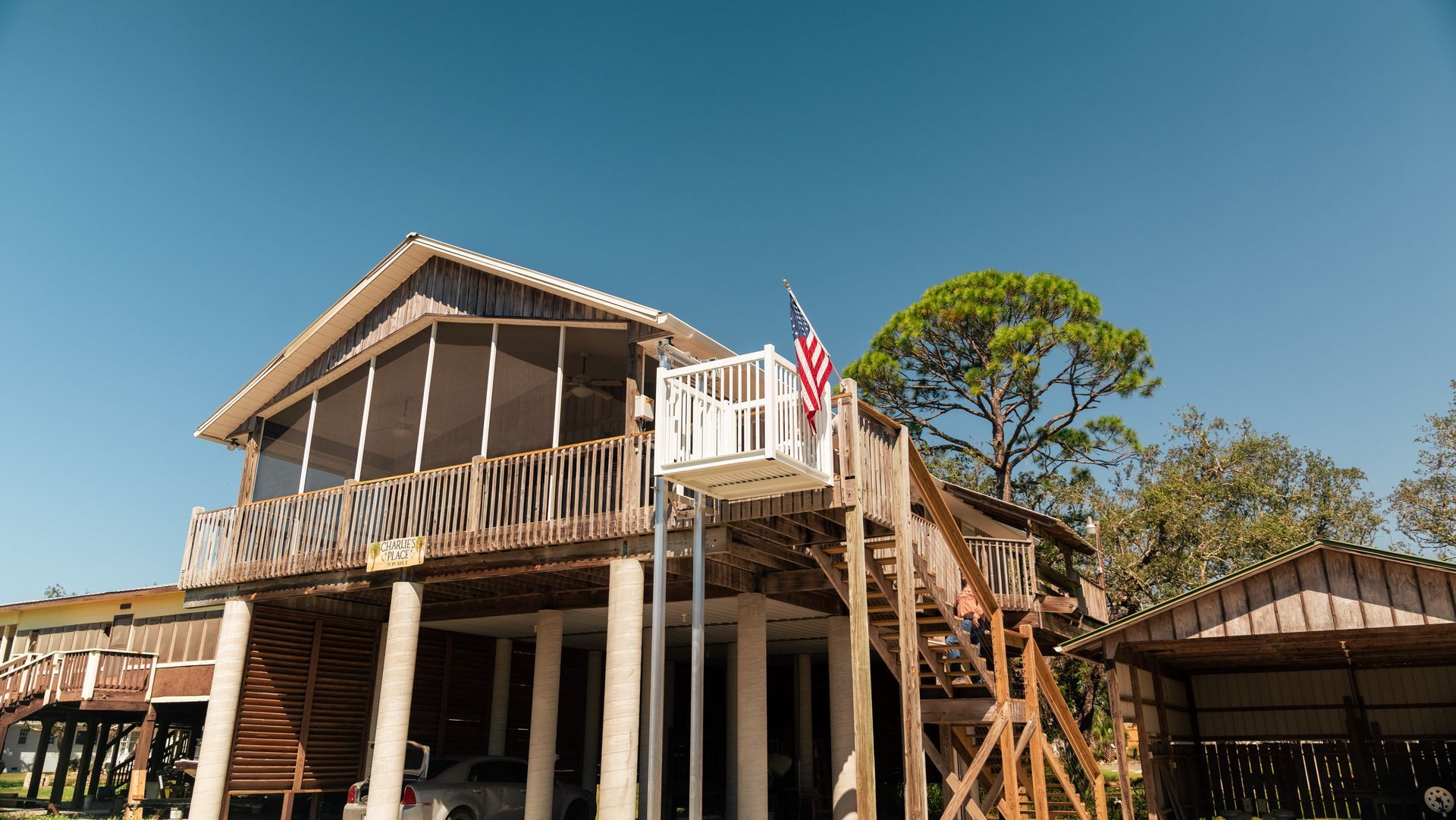 A wooden stilt house with a platform lift, American flag, and a screened-in porch under a blue sky.