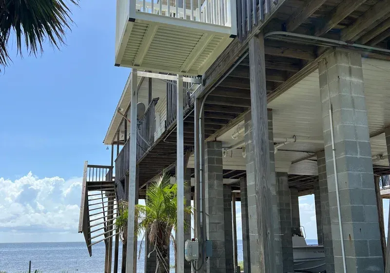 Elevated coastal home on concrete pillars, wooden decks and stairs, blue sky.
