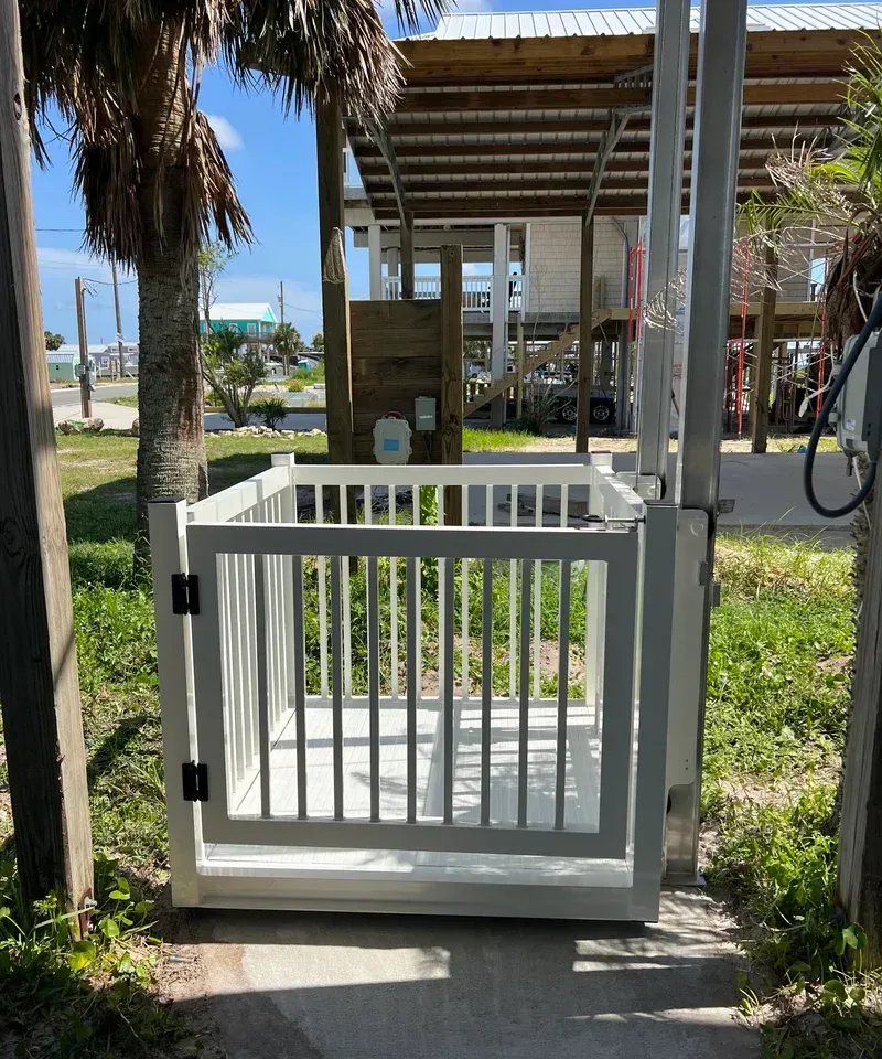Outdoor wheelchair lift, white, with gated platform, next to a building on pilings.