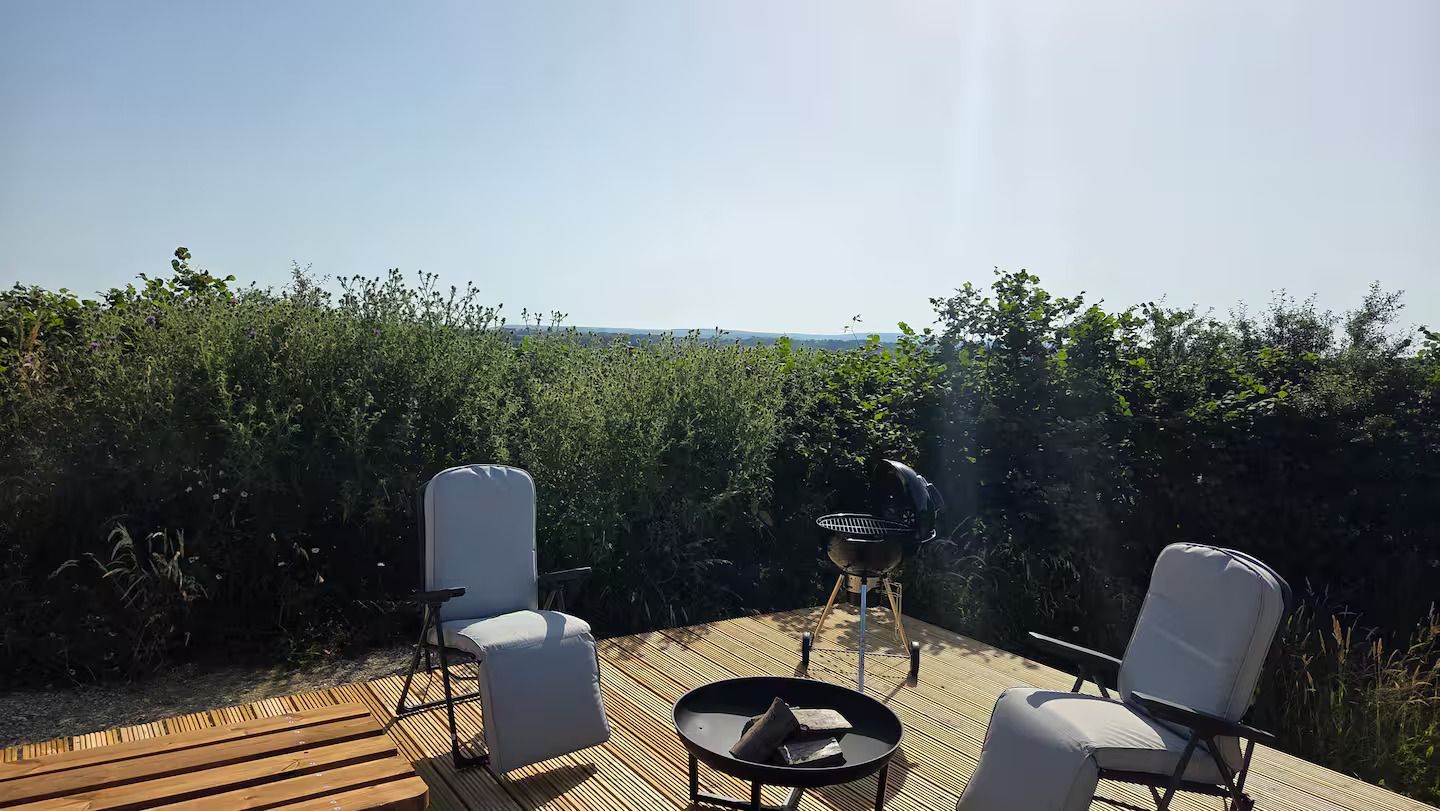 Patio seating area with white chairs, a round fire pit table, and a hedge under bright sunlight.