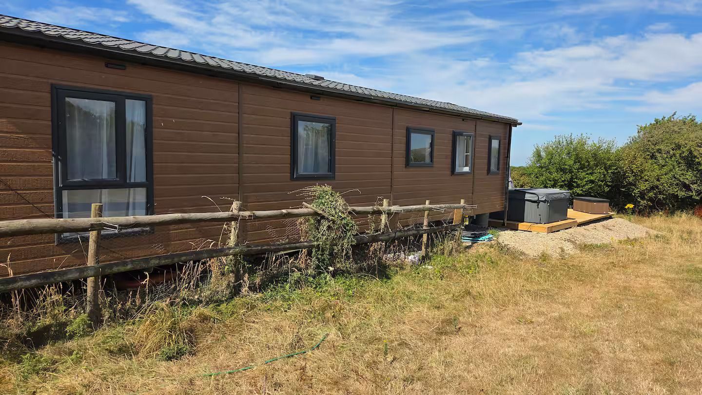 Long brown house with dark windows beside a dry grassy yard and bushes under a blue sky
