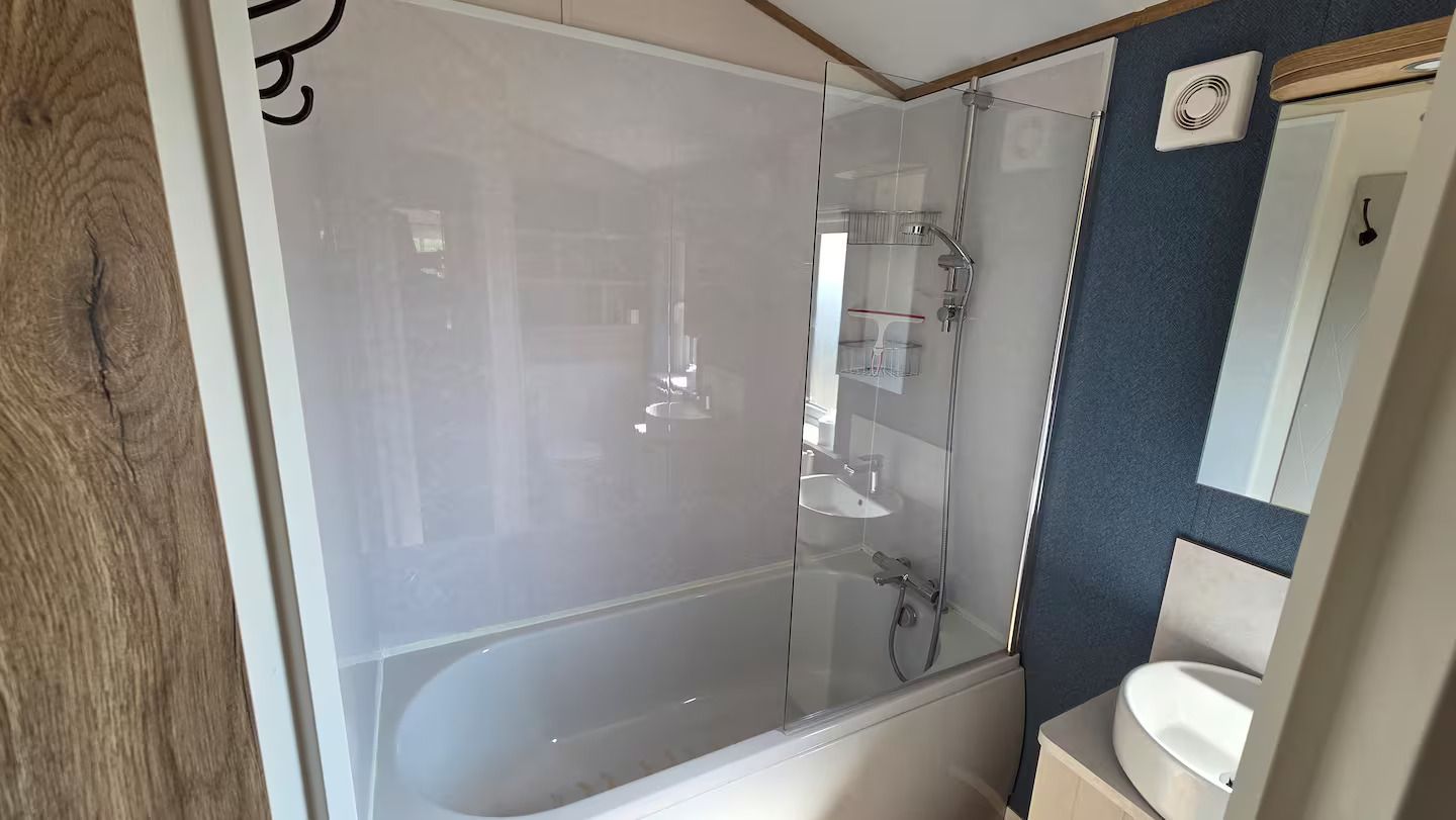 Small bathroom with a white bathtub, glass shower screen, wall-mounted mirror, and sink.