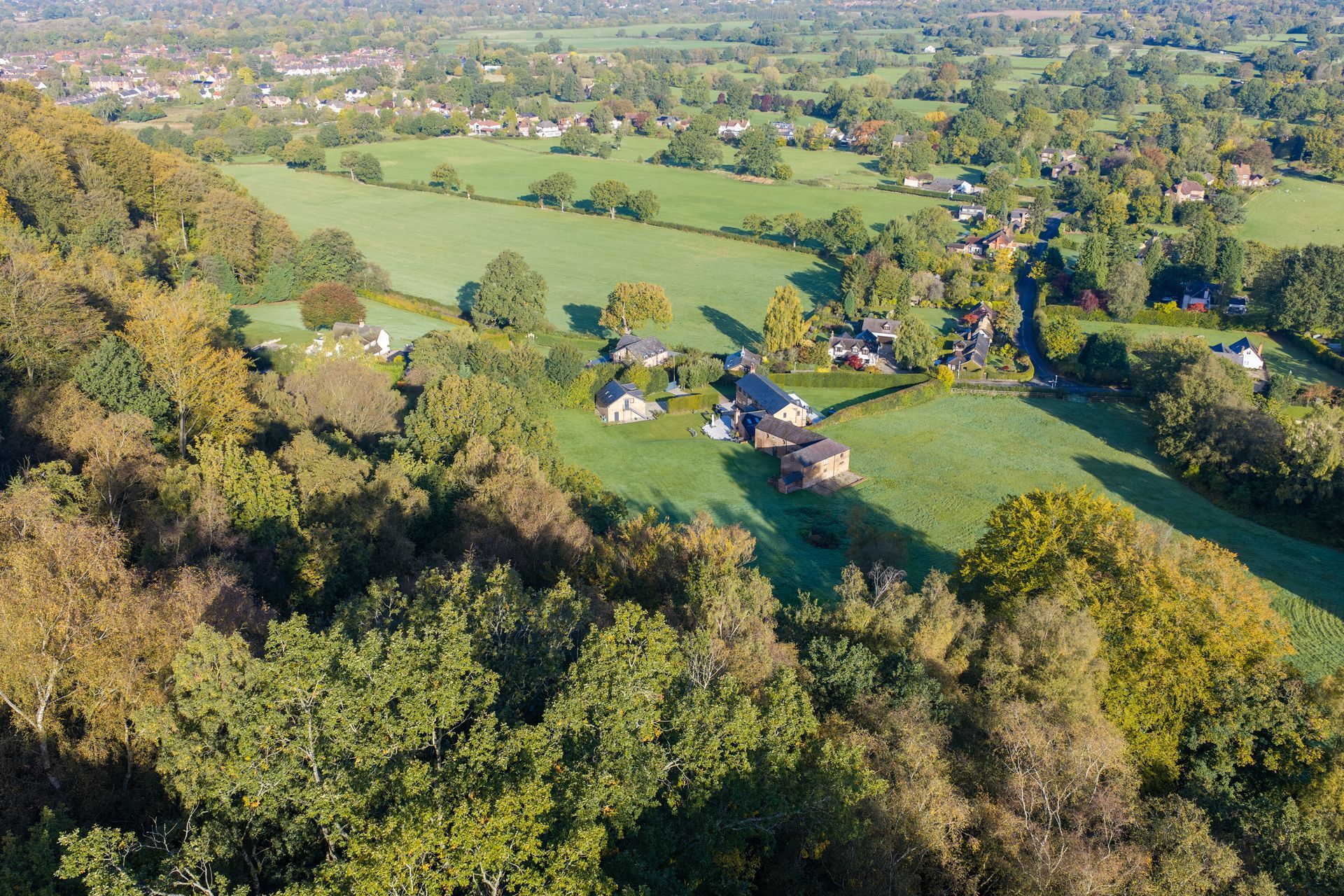 Aerial view of a green countryside with fields, scattered houses, and a wooded hillside.