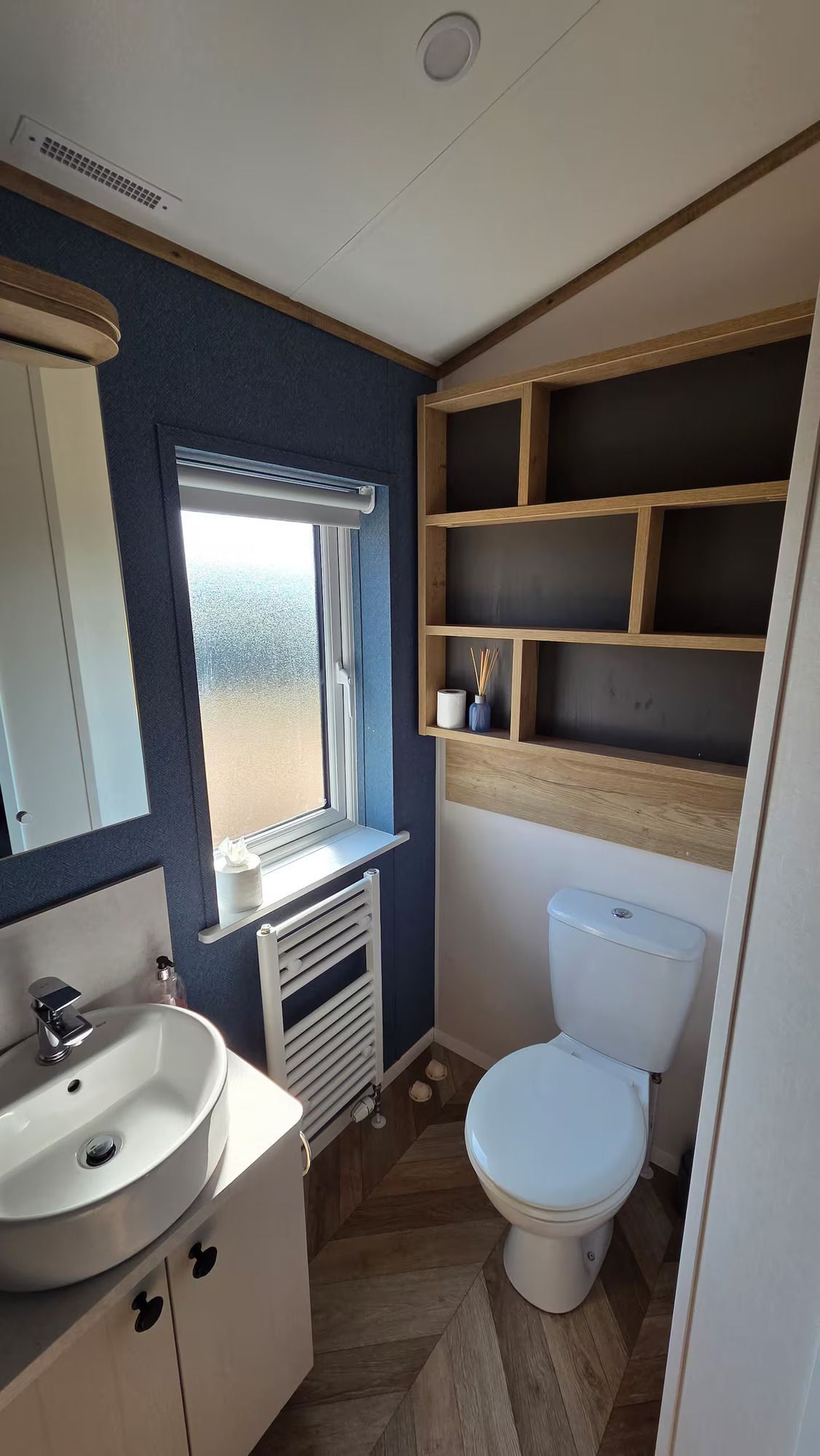 Small bathroom with white toilet, sink, window, and built-in wooden shelves.