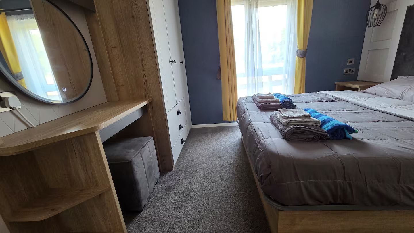 Bedroom with two neatly made beds, a vanity mirror, wardrobe, and daylight through a window.