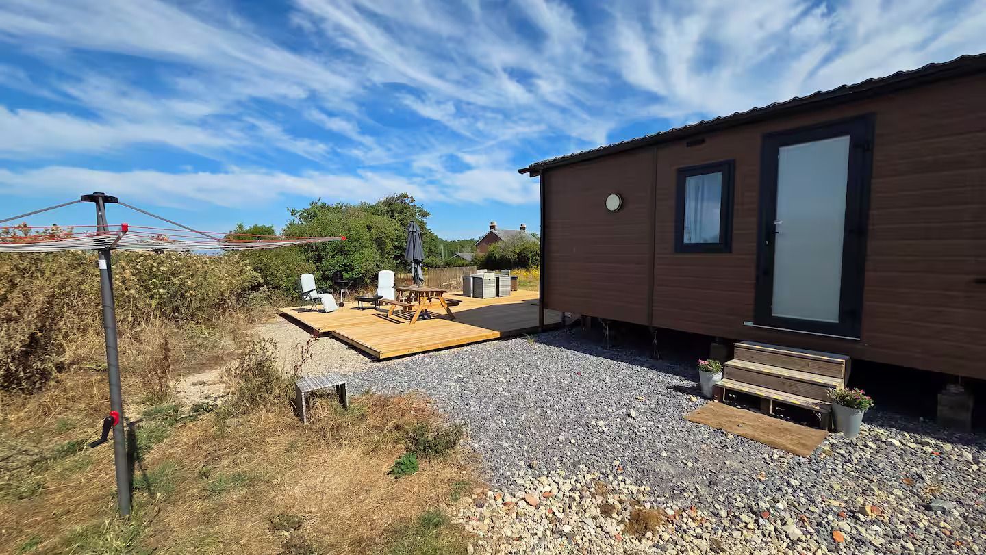 Small brown cabin beside a gravel yard, with a wooden deck, hedge, and blue sky.