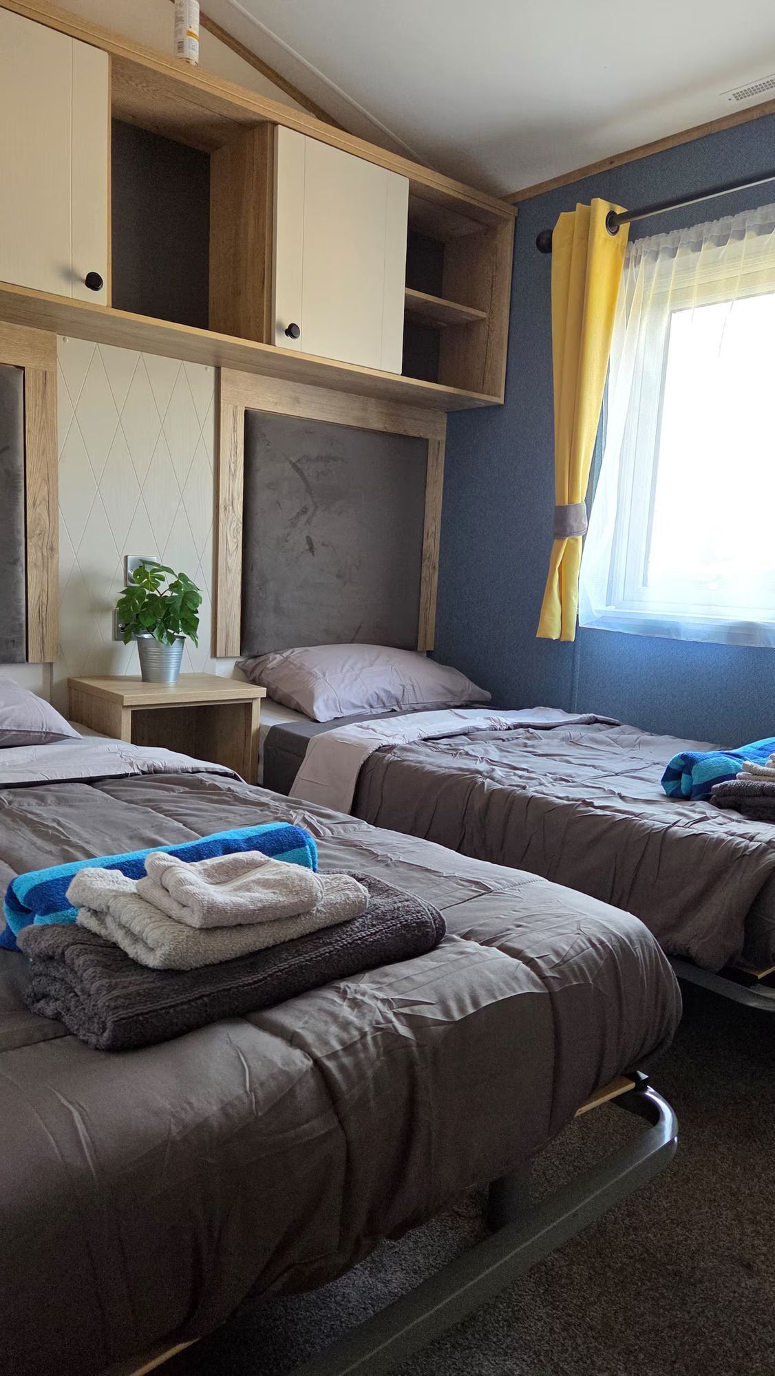 Small bedroom with two single beds, built-in cabinets, blue walls, and a bright window with yellow curtains.