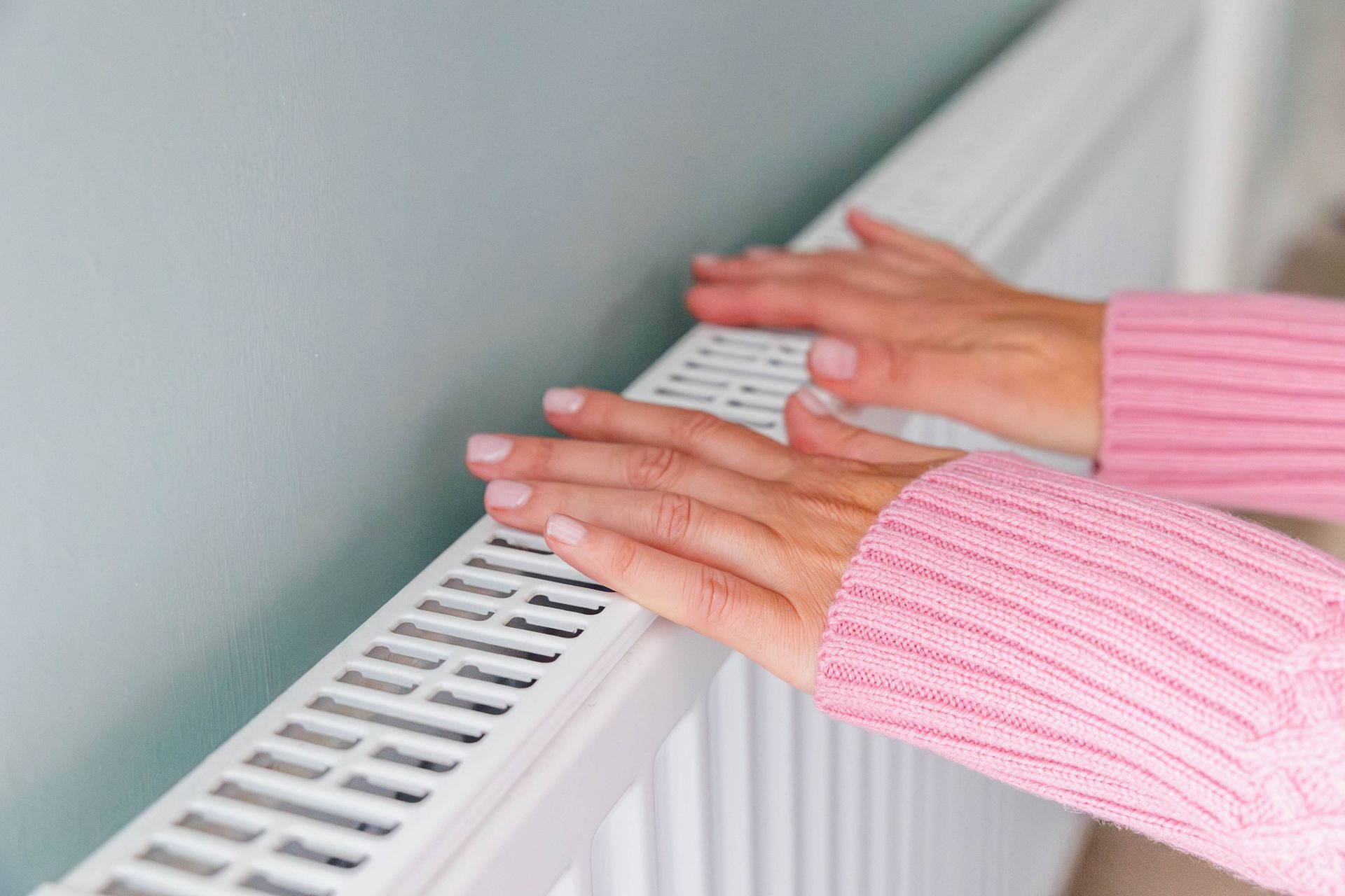 A woman in a pink sweater is touching a radiator with her hands.