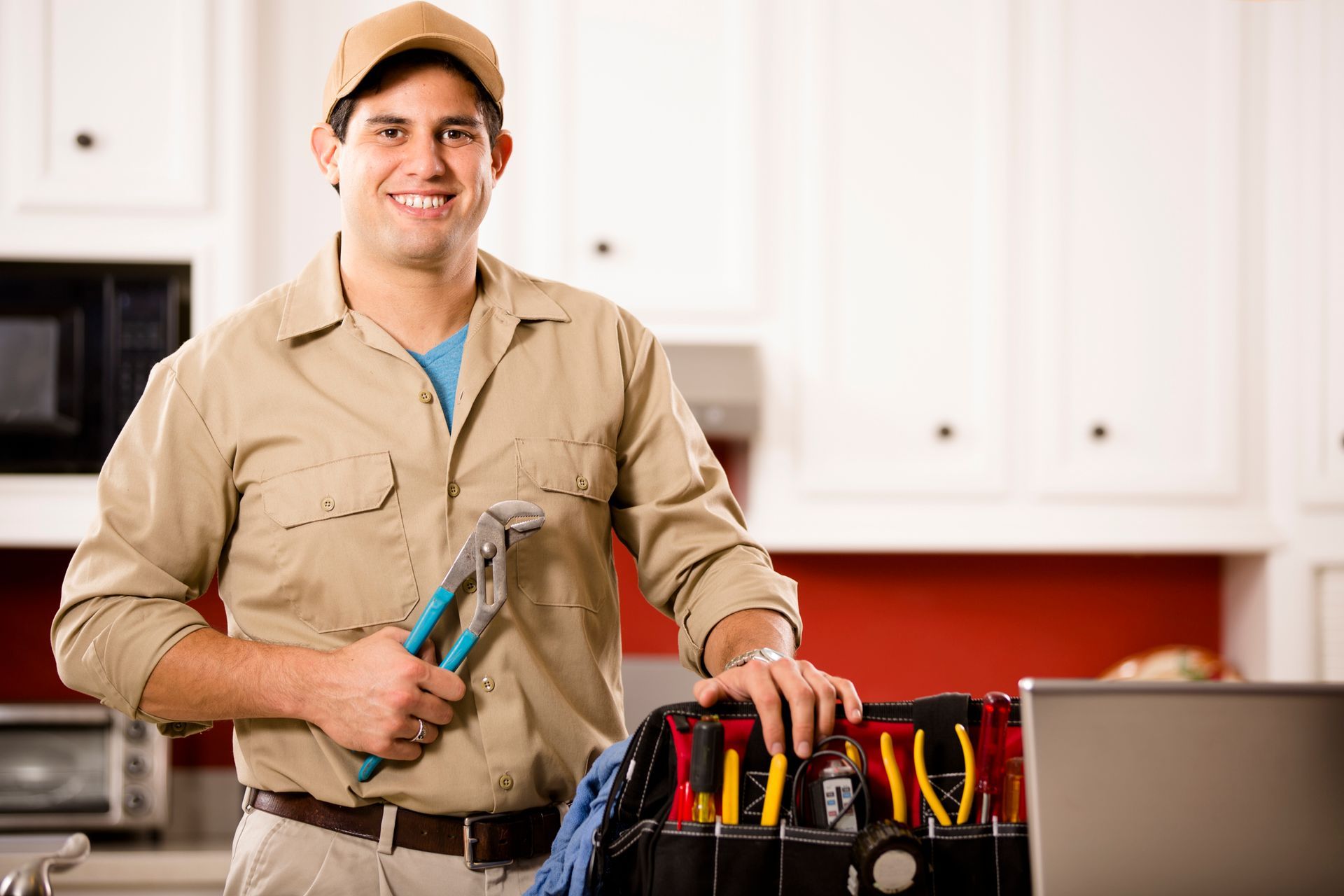 A man is standing in a kitchen holding a wrench and a toolbox. A man is standing in a kitchen holding a wrench and a toolbox.