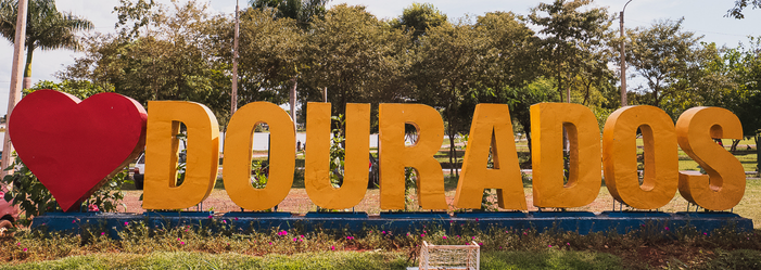 Placa turística com a frase “Amo Dourados” em área arborizada da cidade de Dourados, Mato Grosso do Sul.