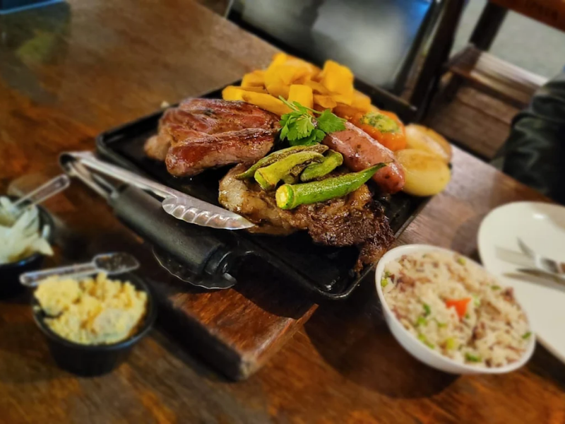 Prato com carnes grelhadas, linguiça, legumes, arroz e acompanhamentos servido à mesa.