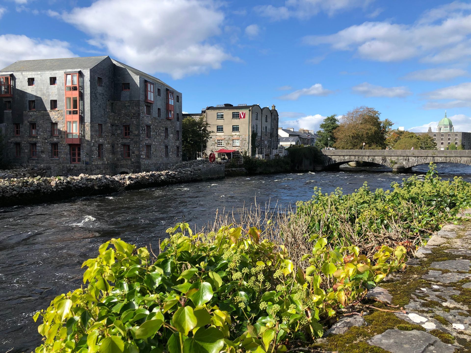 River with stone buildings on the banks, bridge in the background, blue sky with clouds.