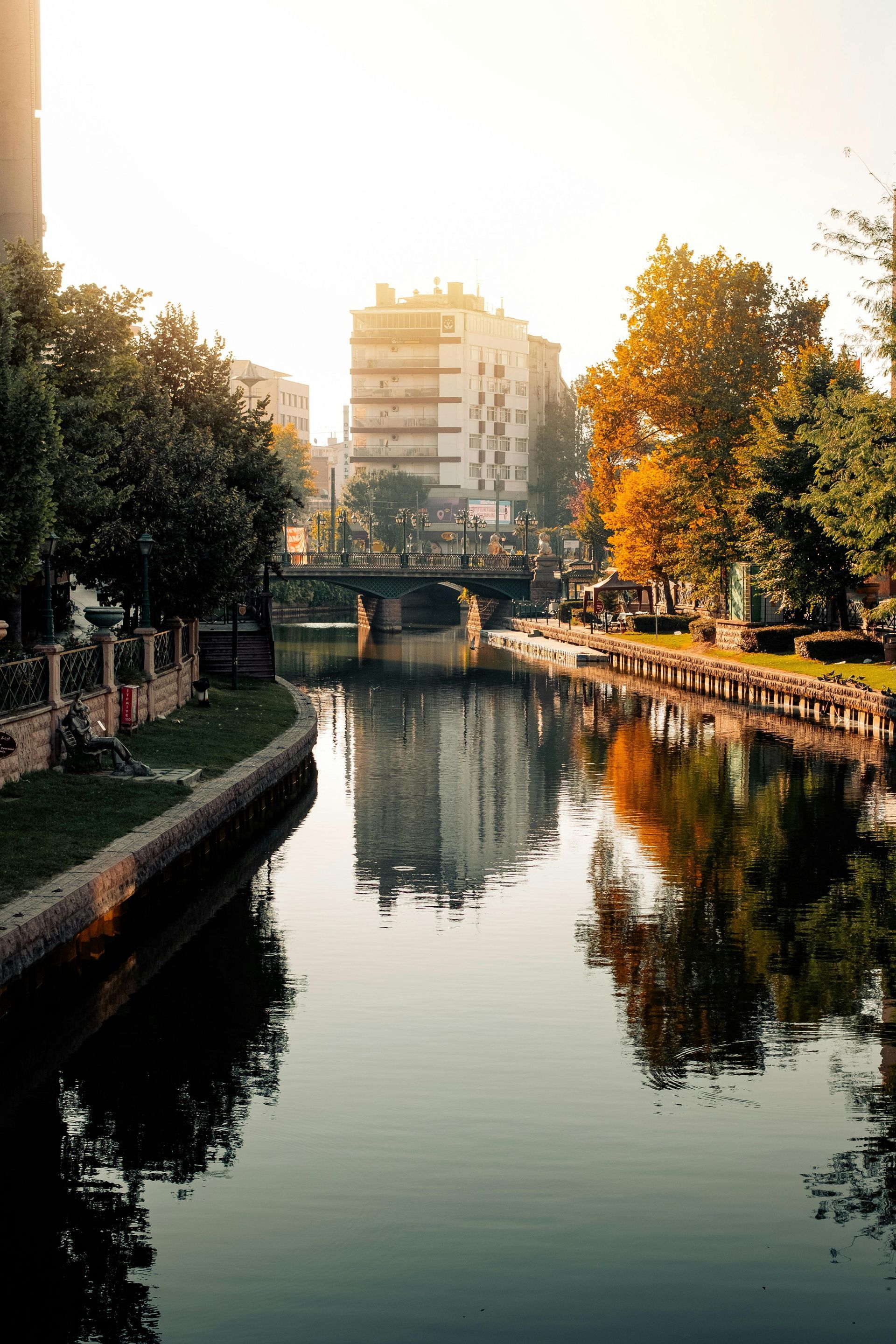 Canal with buildings, trees, and bridge reflecting on the water, bathed in sunlight.