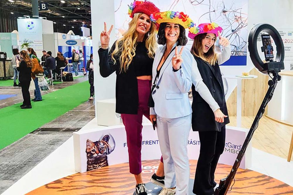 Three people in colorful wigs posing at a trade show booth with a ring light and promotional display