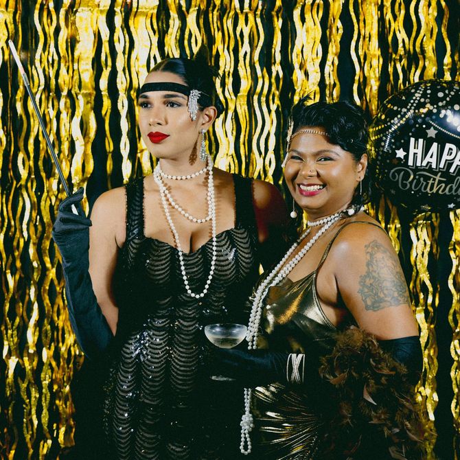 Two people posing at a birthday party in black outfits against a gold streamers backdrop