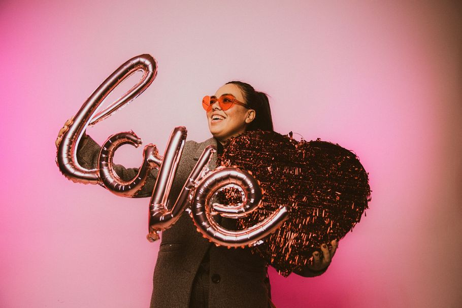 Person holding a large metallic “Love” balloon against a pink background, smiling in sunglasses