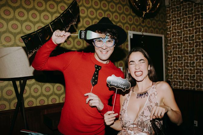 Two people in costume posing at a party, one in a red sweater and the other holding a drink and smiling.
