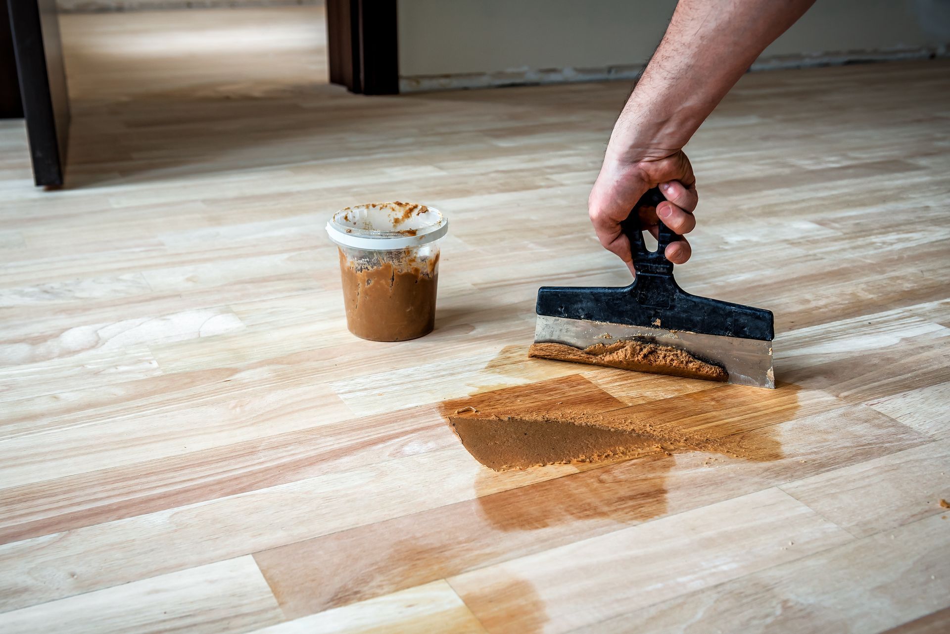 Une main utilise un grattoir pour enlever une tache brune renversée sur un parquet clair, à côté d'une tasse de teinture.