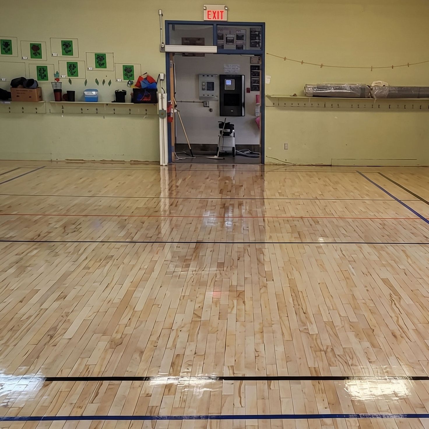 Sol vide d'un gymnase faisant face à une porte ouverte sous un panneau SORTIE, avec des étagères le long des murs.