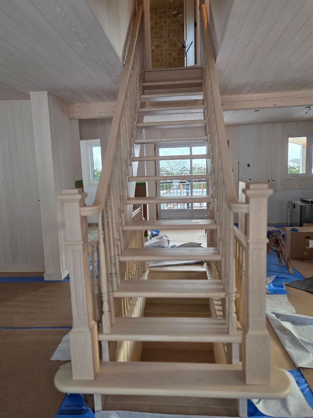 Escalier en bois en construction dans une pièce lumineuse, avec des rampes inachevées et des outils à proximité