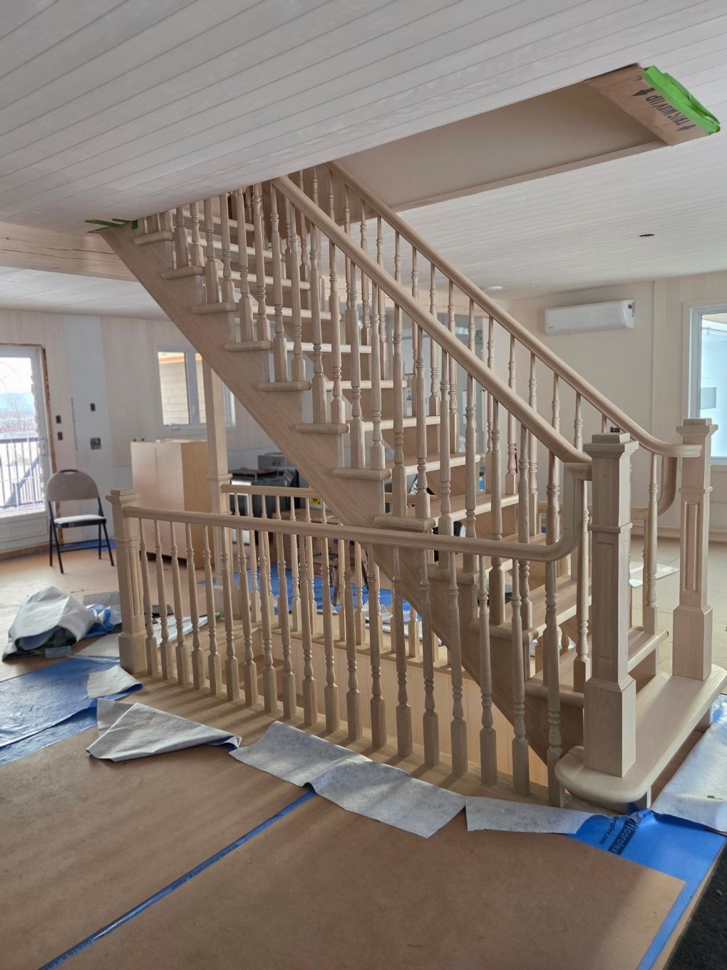 Escalier en bois en construction dans une pièce lumineuse, avec une bâche de protection au sol et du ruban adhésif bleu.