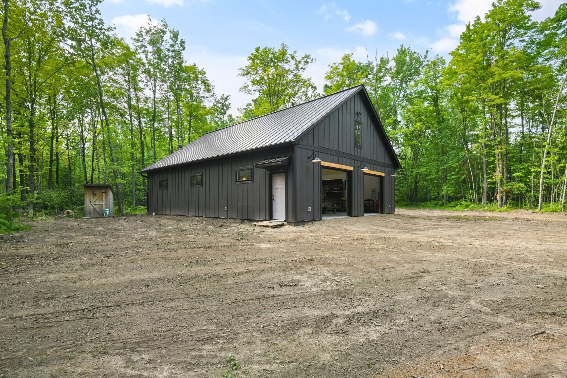 Dark gray barn-style building in a dirt clearing, surrounded by green trees under a blue sky.