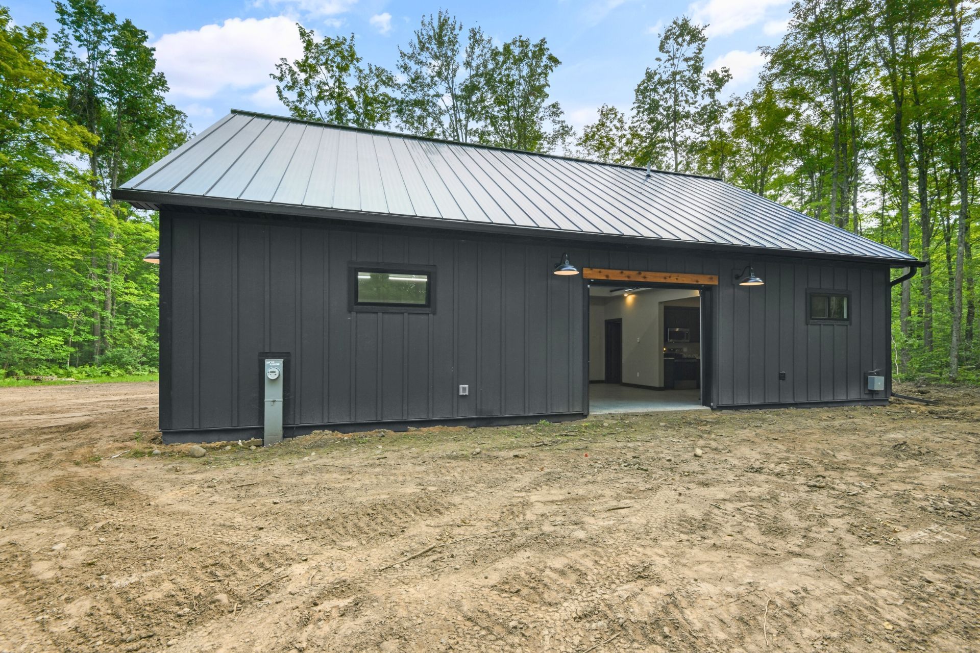 Dark gray metal barn-style building with a sloped roof, set on a dirt lot surrounded by trees.