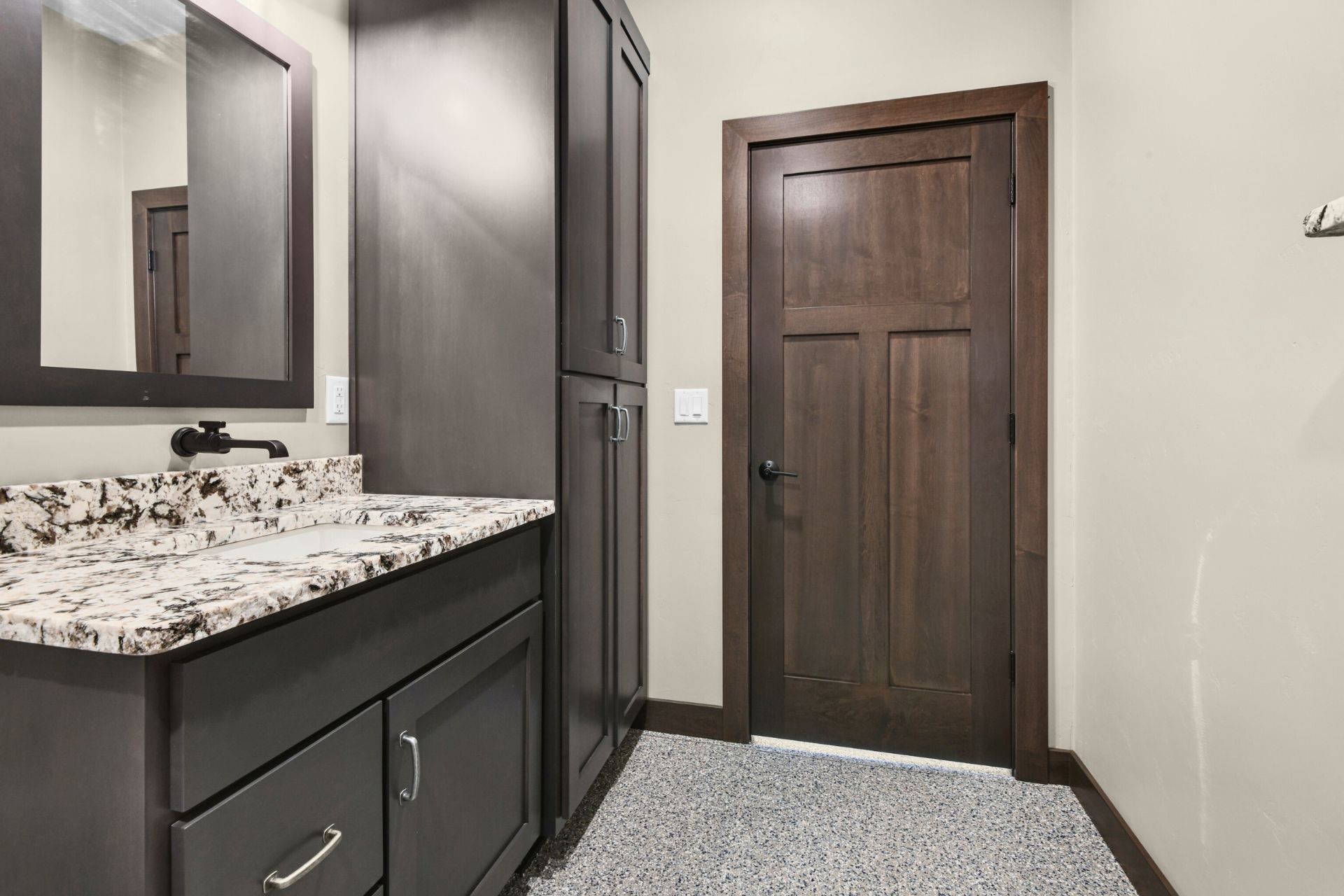 Bathroom vanity with dark cabinets, granite countertop, mirror, and a wooden door on the right.