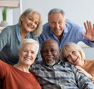 Five smiling adults leaning together on a couch, waving at the camera.