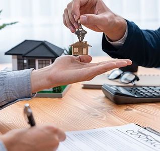 Hands exchanging house keys over a desk with contract, pen, laptop, and a model house.