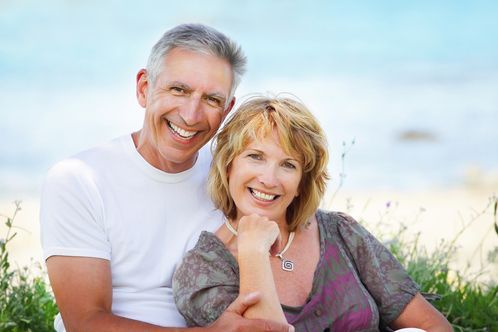Smiling couple sitting on a beach, with the man in a white shirt and the woman in a sheer top.