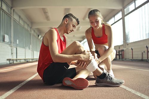 Two athletes in red sportswear sit on a track, wrapping an injured ankle.