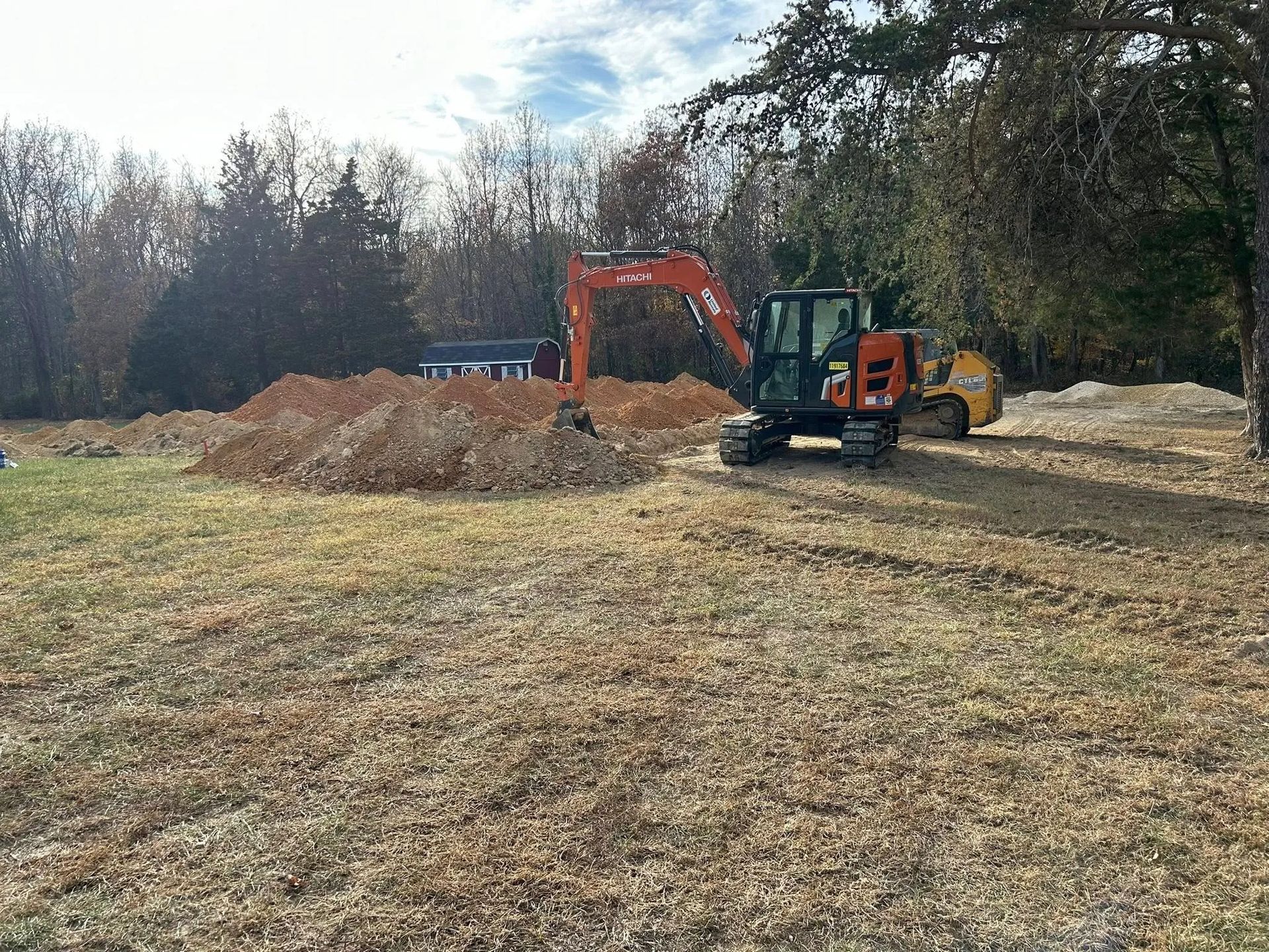 Excavator and skid steer moving dirt on a grassy construction site with trees in the background