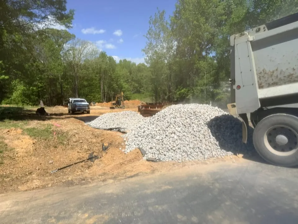 Gravel pile beside a dump truck on a sunny dirt road, with trees and a parked vehicle in the background