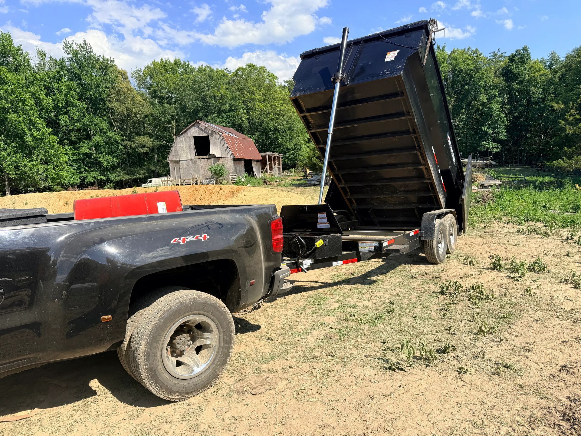 Black dump truck with raised bed parked on a dirt lot near a red barn and trees.