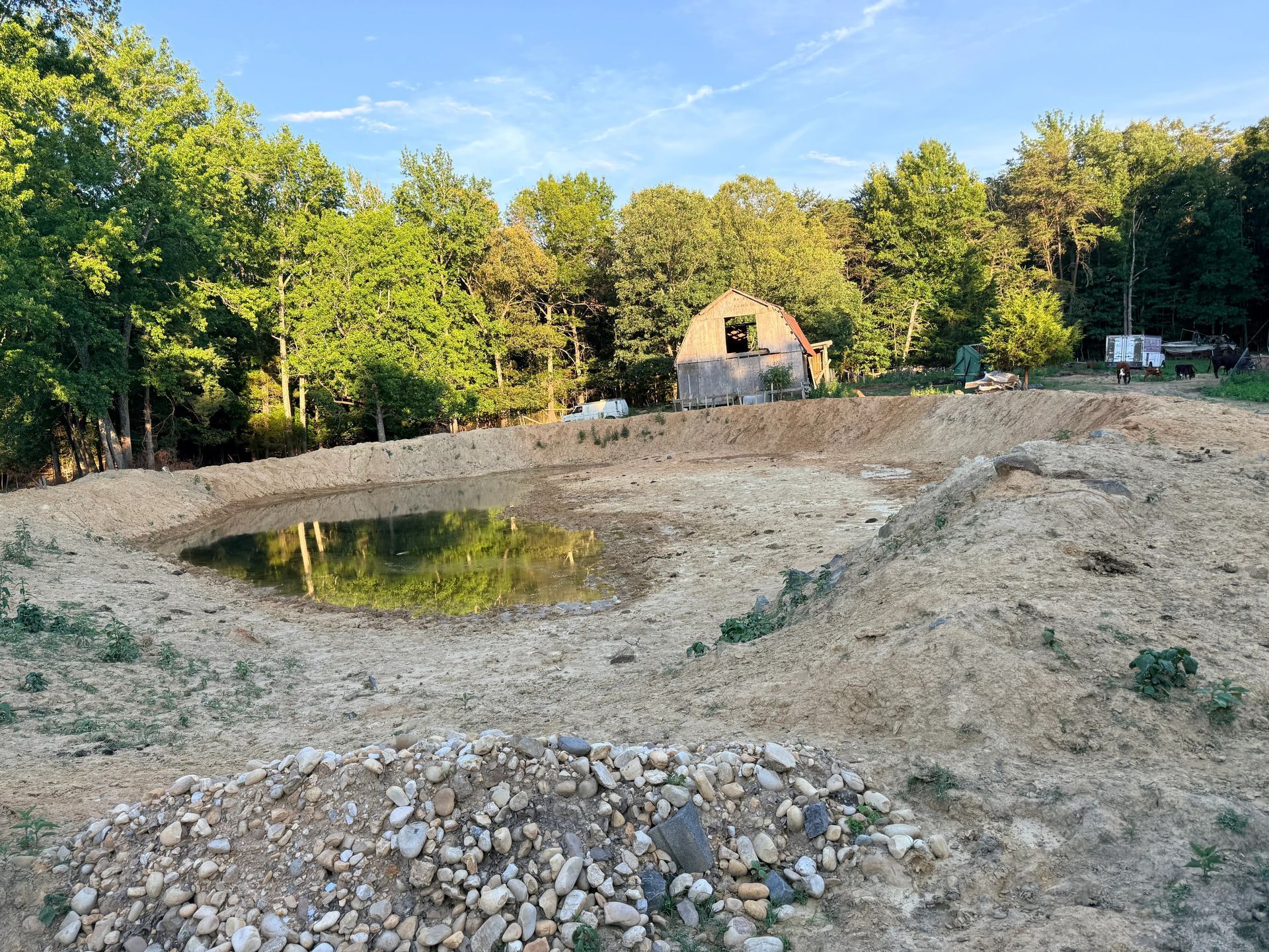 A muddy excavation site with a shallow pond, dirt mounds, and trees surrounding a small building in the distance