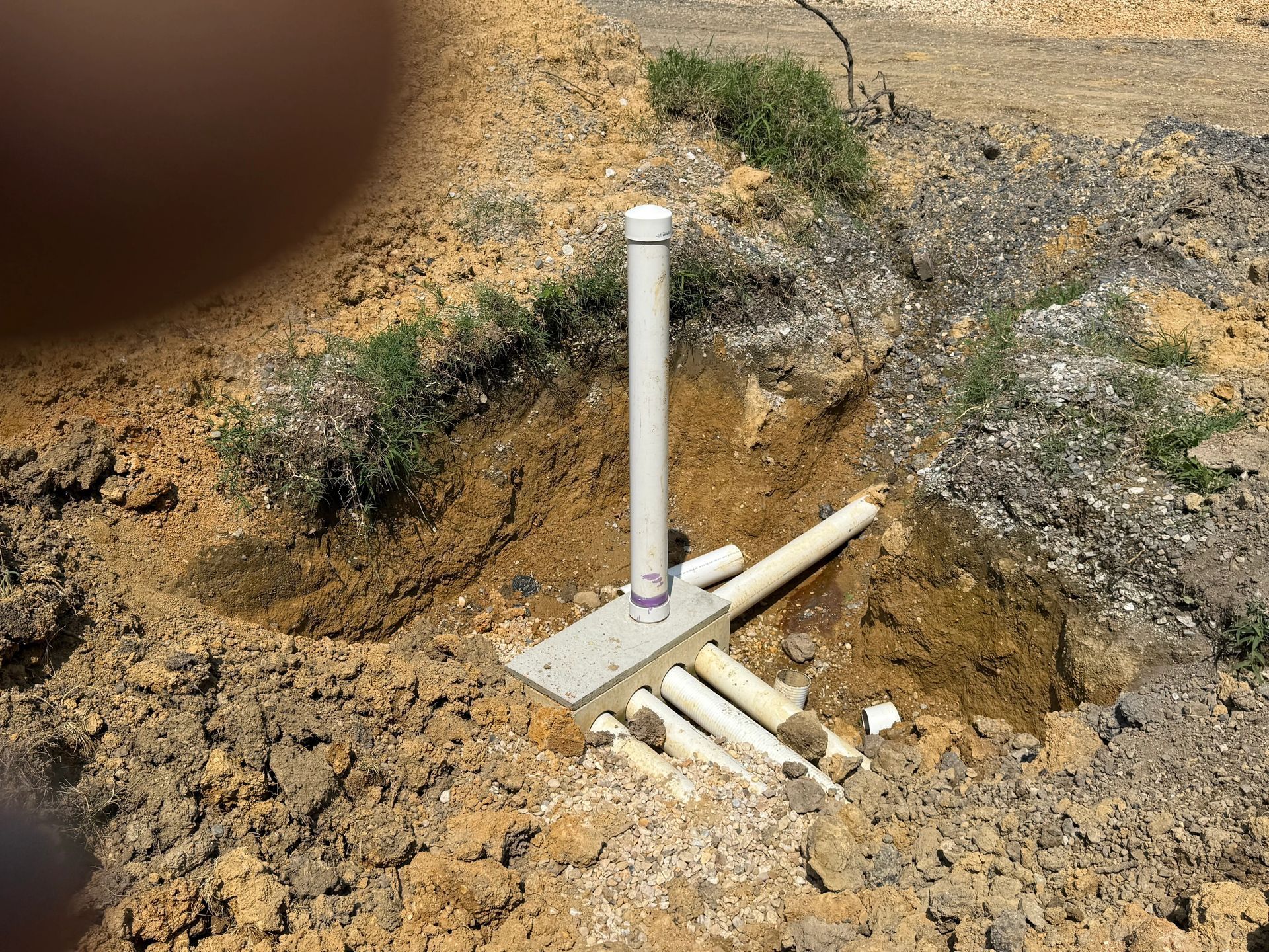 White PVC pipes and fittings protruding from a dirt hole in rocky ground.