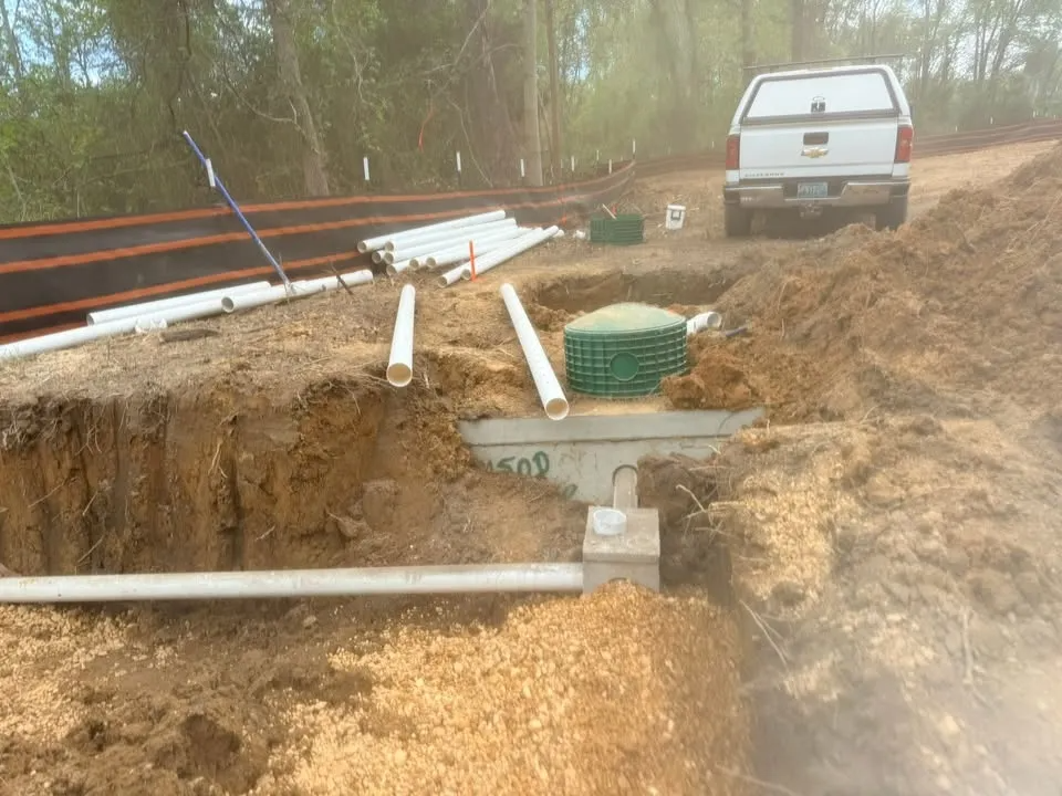 Construction trench with exposed pipes and green utility box beside a white pickup truck on a dirt site