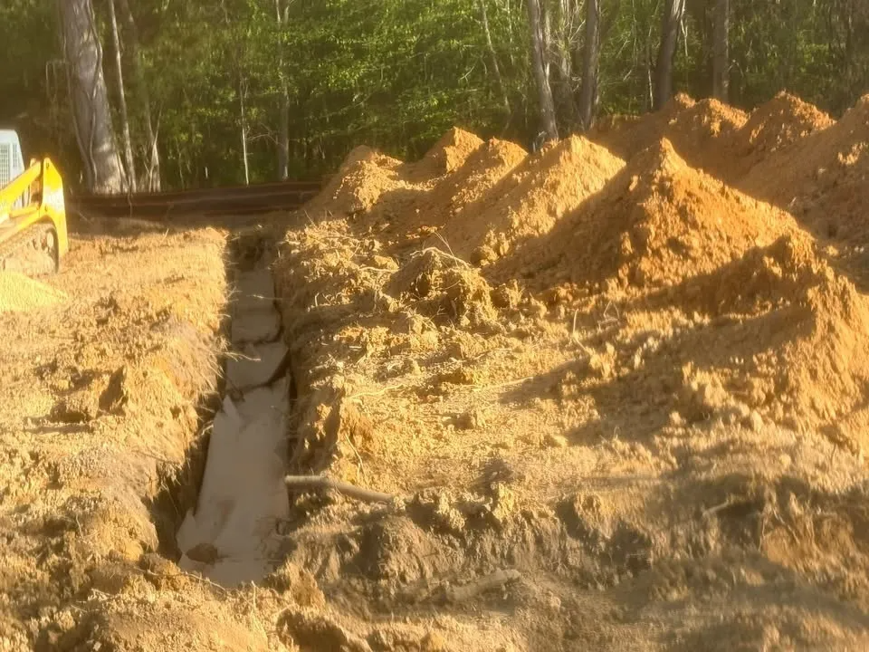Construction trench beside dirt piles in a wooded area, with part of a yellow excavator visible.