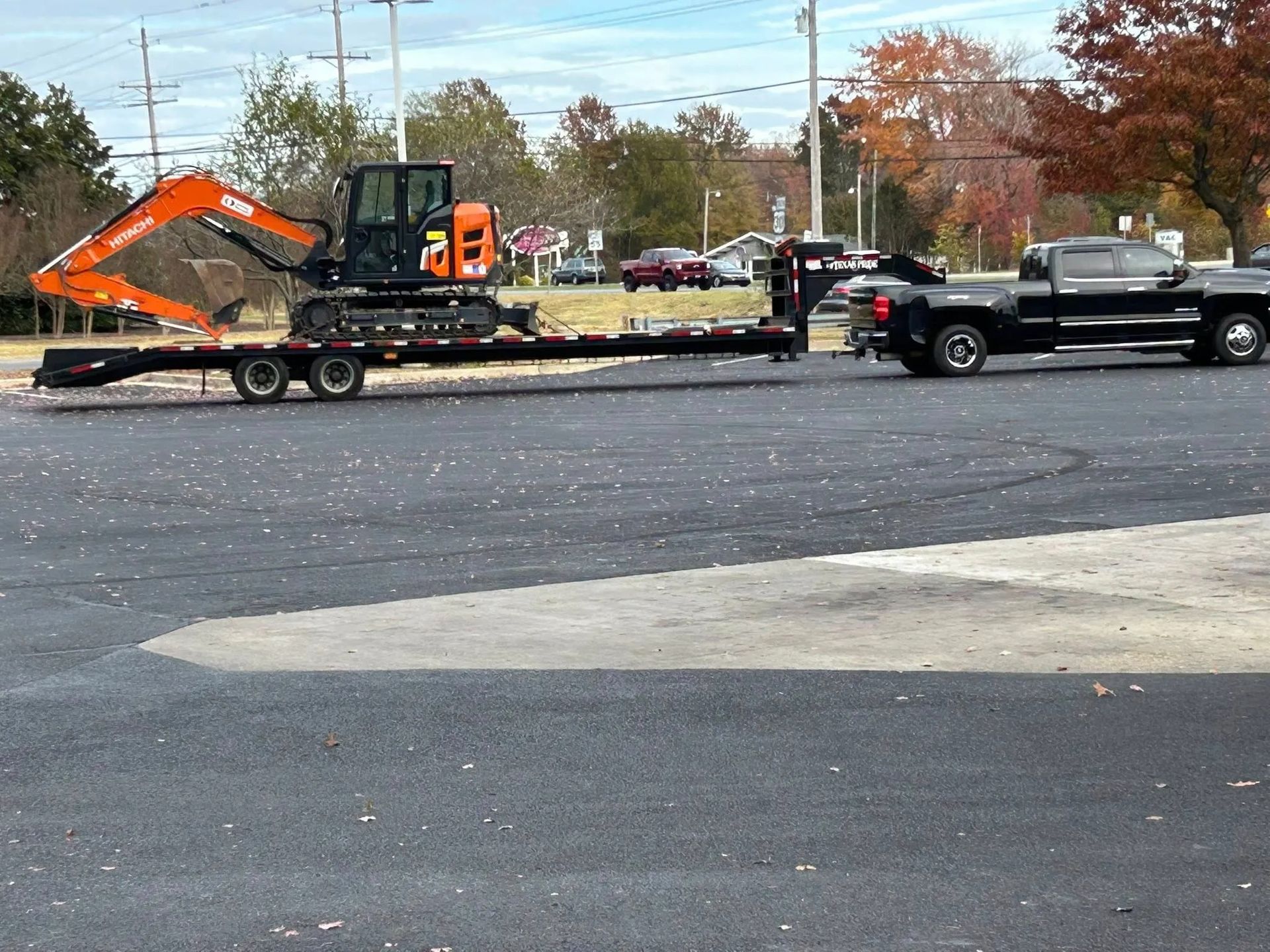 Orange excavator on a flatbed trailer being towed by a black pickup in a parking lot