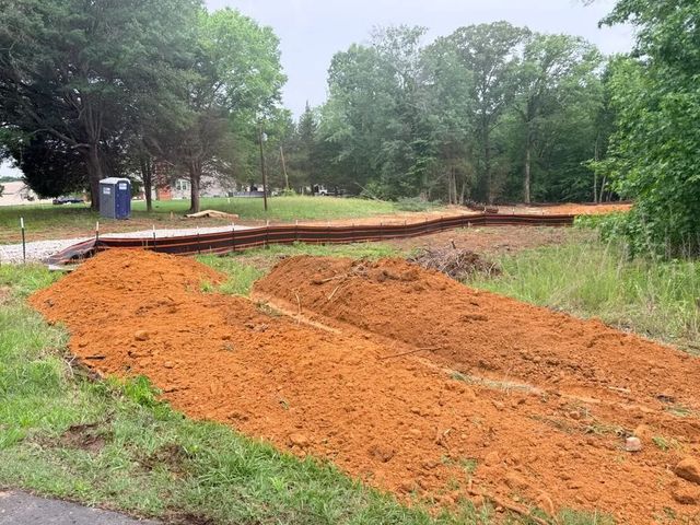 Red dirt path under construction in a grassy park with trees and a portable toilet nearby