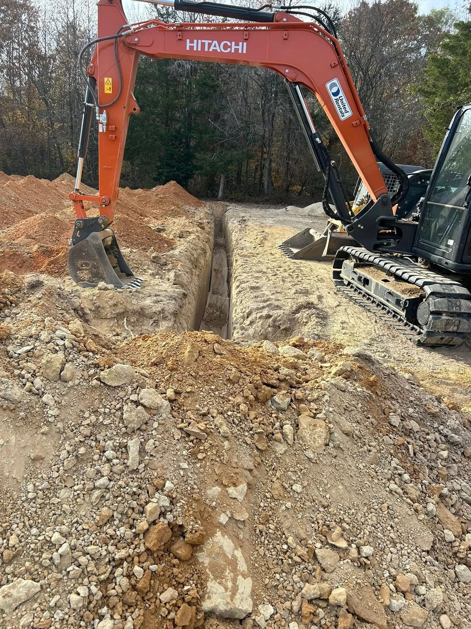 Orange excavator digging a trench in a muddy construction site beside a wooded area