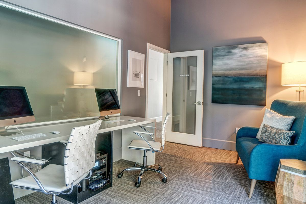 Interior community workspace with two iMacs on a long white desk, white chairs, a frosted glass divider, and a blue sofa.