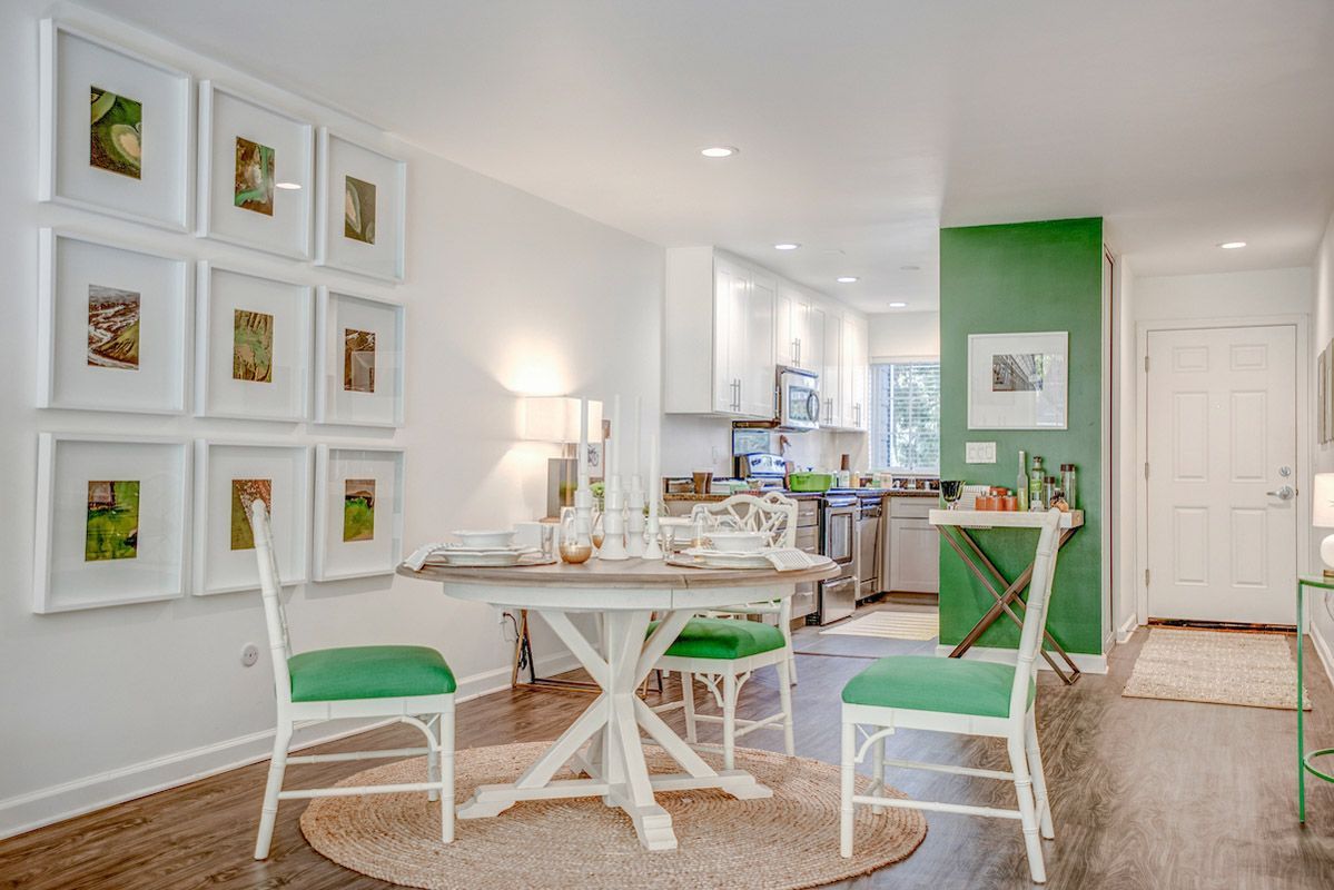 Open dining area in a modern apartment: round table, white chairs with green cushions, kitchen in background.