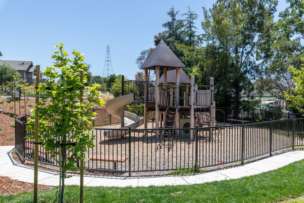 Wooden playground structure with slide, climbing elements, and a surrounding black metal fence in a sunny courtyard.