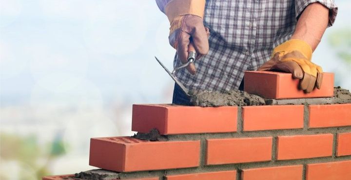 Worker laying red bricks on a wall with mortar and trowel, wearing gloves outdoors