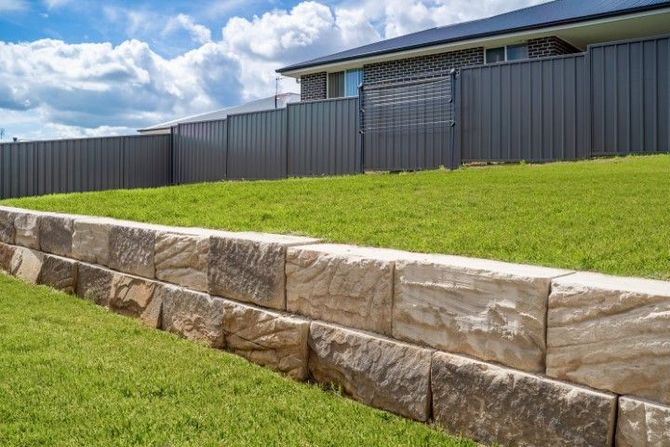 Stone retaining wall beside a green sloped lawn and gray fence under a cloudy blue sky