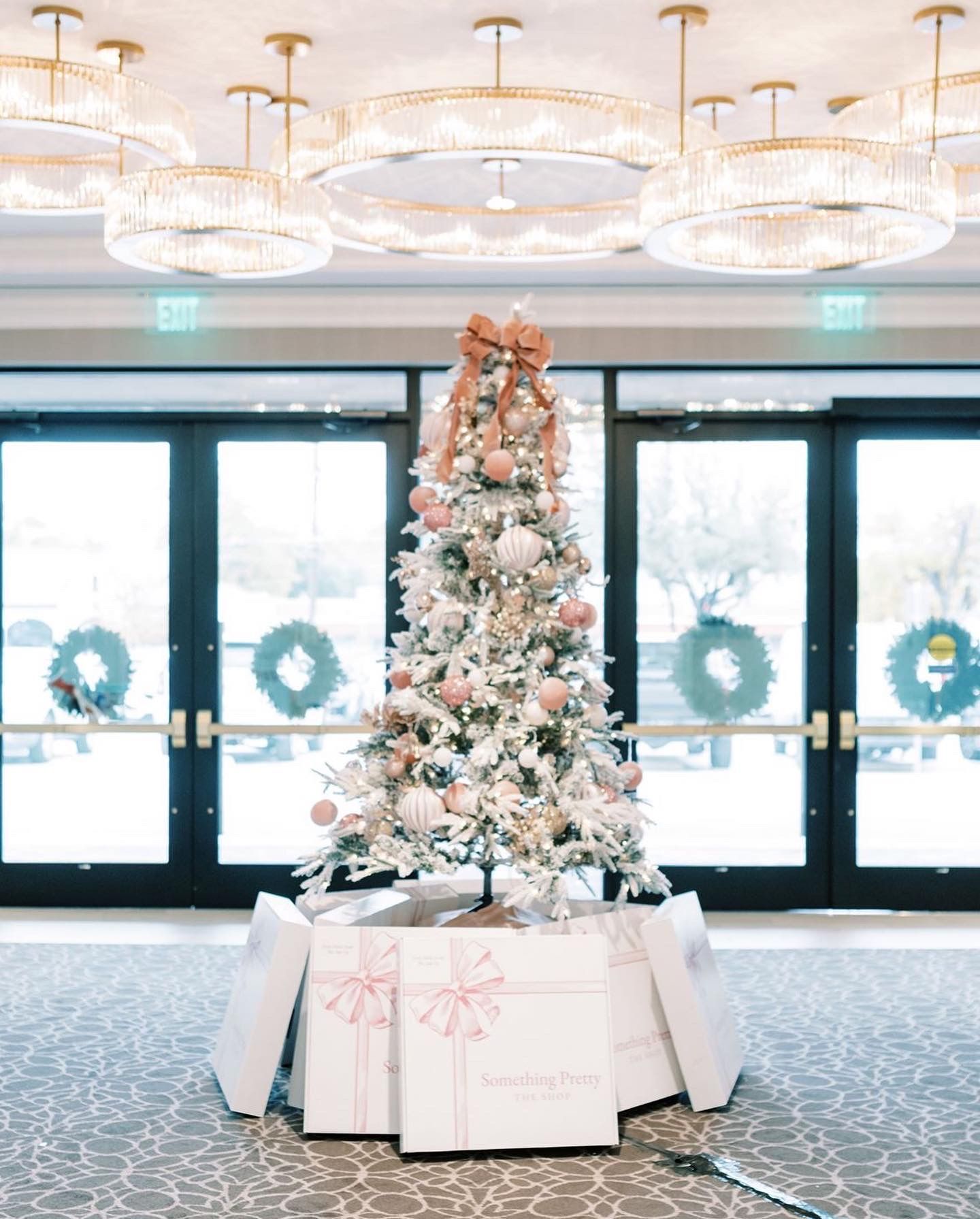 Christmas tree with pink ornaments and gifts, indoors. Large glass doors and round chandeliers in background.